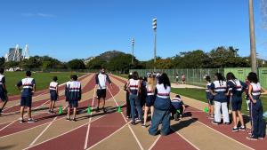 Foto dos alunos em pé na pista de atletismo, ouvido a explicação de um professor