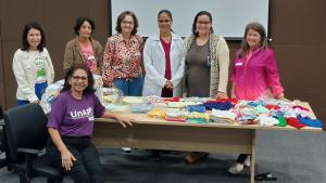 Foto da equipe do hospital com as crocheteiras, em volta de uma mesa onde estão as peças doadas
