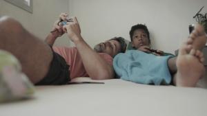 Cena de Lar, na qual aparecem um rapaz e um menino jogando videogame deitados em uma cama